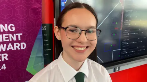 Rebekah smiling at the camera. She is wearing black framed round glasses, a white shirt and green tie.