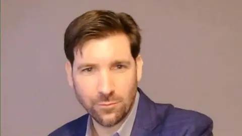 Young man with short brown hair and short beard looks towards camera on a video call. He is wearing a dark blue suit jacket. 