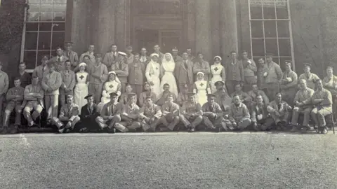 North Lincolnshire Museums A group of about 50 soldiers in their hospital uniforms are standing or sitting in front of the entrance to Normanby Hall. Five nurses are also in the group.
