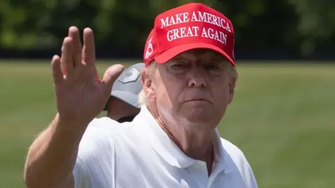 EPA Trump na polu golfowym w czerwonej czapce America Great Again i białej, zbiorowej koszulki golfowej w maju 2023 roku. Ma machanie i patrzy bezpośrednio na kamerę, zielony tor wodny za nim