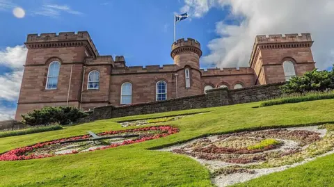 A view of Inverness Castle looking up to it from Castle Street. The castle is a sandstone building with ramparts and turrets. Long windows, curved at the top line the walls and a Saltire flies from a flag pole in a round turret. Below the castle is a large area of grass with floral displays, including a clock. It is a bright sunny day.
