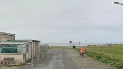 A road leading to a sandy area and the sea at Walney. There is a pavement on which a person in a pink coat and dark trousers is walking a dog. There is long grass on the right. There is also roadside no entry sign. There is a small brick building with a flat roof and temporary fencing around it on the left.