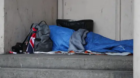 The outline of a person in a blue sleeping bag on the concrete steps of a doorway. They have a grey backpack with a union flag tied to it next to them and a larger black cube bag behind them