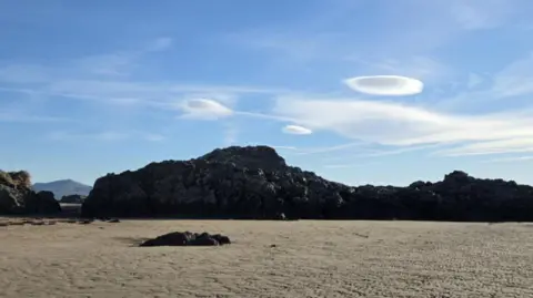 Emily Camp The picture shows a mountain on the beach, there is sand and a blue sky with 3 flat (UFO) clouds in the skyline.