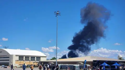 Black smoke rises into a blue sky over two buildings at a US military base.