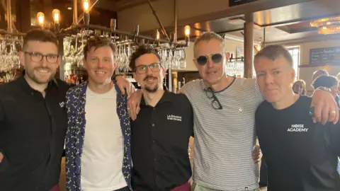 Five men stand in front of a bar, all smiling at the camera with their arms around each other's shoulders.