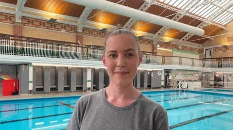 Sally Fairfax/BBC A young woman is standing in front of a swimming pool. She is smiling and wearing a T- shirt that says "Synergy Body and Mind" on the chest.