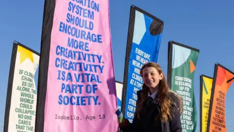 A young girl in a school uniform standing in front of colourful flags displaying inspirational messages. The pink flag closest to her reads, "The education system should encourage more creativity. Creativity is a vital part of society." The sky is clear and blue.