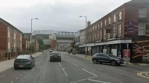 A wide road with a row of shops on one side and a modern two-storey block of flats in the left hand side. Anfield Stadium is in the distance and three cars are travelling towards the ground and one car is turning into a side road.