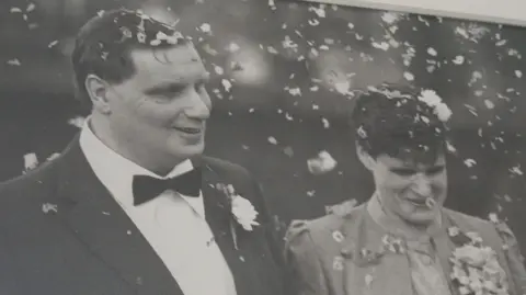 Lynne and Trevor Man with dark suit, white shirt, bow tie and flower on his pocket and with short hair and woman with light jacket and light top with short hair. There is confetti in the air as they both smile on their wedding day.