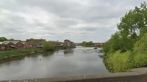Google A canal, pictured from a bridge above. Trees and industrial buildings can be seen flanking the water.