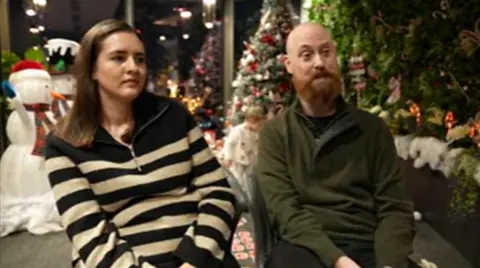 A woman with long brown hair, wearing a navy and cream-striped top and a man with a red beard and wearing a green top, sit in front of Christmas decorations.