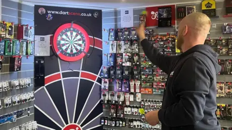 Ryan Clarke throwing a dart in his shop surrounded by darts equipment on shelves.