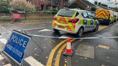 Stephen Huntley/BBC A sign that says Police - Road Closed stands at the junction in front of the gutted house. There are three police vehicles parked up behind it, on the opposite side of the road to the house.
