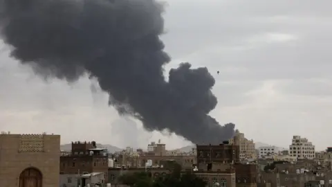 Thick grey smoke billows after the air strike in the distance. In the foreground are lots of buildings
