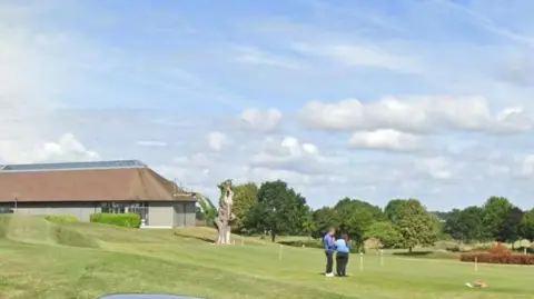 Google A golf course, with two people dressed in blue and black standing near a hole. There are trees in the background and a clubhouse to the left.