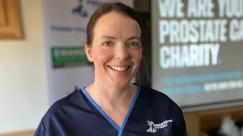 Nurse Amy Griffiths smiling, wearing navy scrubs with Prostate Cymru logo. She has brown hair and blue eyes.