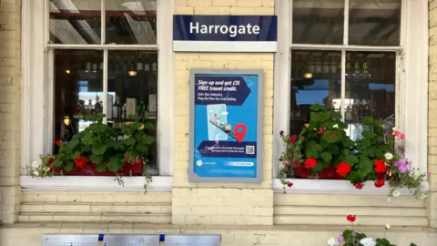 Seb Cheer/BBC A metal bench with blue accents sits beneath a blue “Harrogate” station sign, flanked by two large windows decorated with flower boxes of red, white and purple blooms. A travel‑promotion poster hangs between the windows, showing a smartphone map and a QR code.