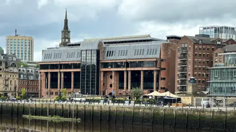 An imposing court building made from red stone and columns with large dark windows, on a river front wiht buildings either side
