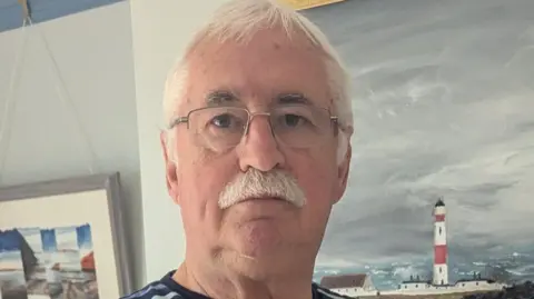 Derek Cunningham A man with short white hair and a white moustache, wearing glasses, stands in front of a white wall with a painting of a lighthouse and the sea on it.
