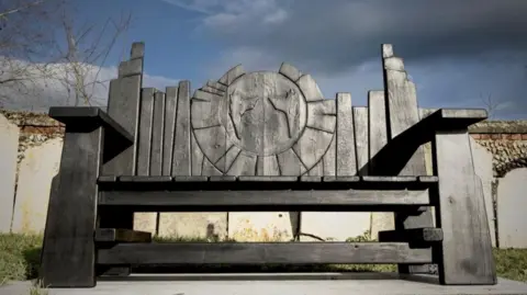 The Prodigy/Facebook A Gothic-style wooden bench in a churchyard. It is dark, possibly black. There is a design etched into the back rest.