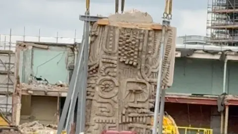 A close-up of the Aztec-style mural being lifted up by metal cranes.