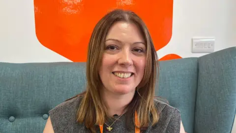 A woman wearing a grey top smiling at the camera. She is sat in a blue chair. Behind her is a white wall with a large orange sticker on it