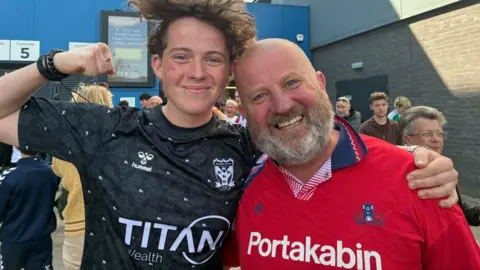 A young man and his dad stand side by side, in York City FC shirts. The young man has curly brown hair, and wears a black shirt with gold detail and the older man has a bald head, a short grey beard and wears a red shirt.