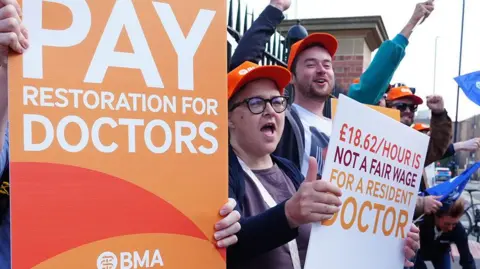PA Media A group of doctors wearing orange BMA hats protest outside iron railings, chanting and raising their fists while holding placards reading “Pay restoration for doctors” and “£18.62/hour is not a fair wage for a resident doctor".