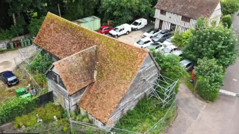 An aerial view of the barn. It is surrounded by fences and scaffolding, and overgrown shrubbery. There are some cars parked between the barn and another half timbered Tudor house. The roof is red with lots of moss growing on it.