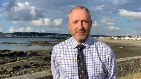 BBC A man wearing a checkered shirt and tie stands with his hands behind his back in front of a beach. 