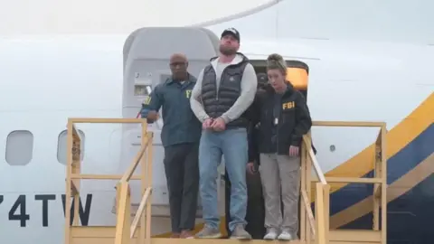 Ryan Wedding stands on steps leading from a plane, with his hands cuffed and surrounded by FBI agents, He wears jeans, a grey hoodie, and a black puffer vest, and has a beard. 
