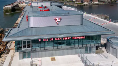 Aerial shot of the front of the ferry terminal, which has Isle of Man Ferry Terminal written across the windows on the second floor and a Manx triskelion on the roof above. There is water either side and red brick building to the right behind it.