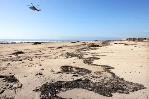 Getty Images A beach with a helicopter overhead. Small pieces of debris, which looks like brown dirt, sits in piles on the sand