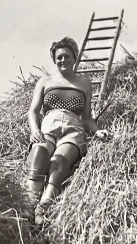 A black and white image of a young Gwen Raggett leaning against a thatched roof. She wears a silk scarf as a strapless top with shorts and leather knee pads on and her hair is wrapped in a headscarf.