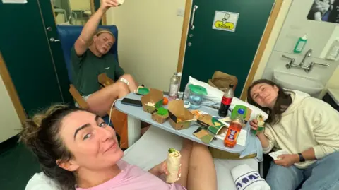 Emma Wassell Emma Wassell in her hospital bed, eating food from the table in front of her while two of her teammates smile for the camera, also with food in hand