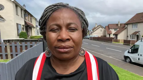 Beverly Simpson has her braided hair tied back and standing in a street in front of houses. She is wearing large black and white hoop earrings, a black T-shirt and red and white scarf.