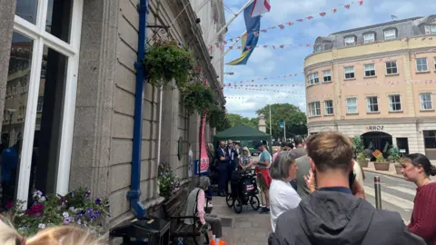 People queue up outside a polling station as they vote in the 2026 Jersey election. It is a sunny day. Flags and bunting are up outside the building.