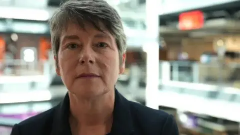 Michelle Morris, looking at the camera. She has short grey hair and is wearing a dark jacket and standing on front of a blurry background of a BBC office.