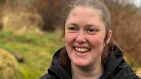 Annabel Rushton from the RSPB. She is mid-conversation and is looking over in the distance. She has brown hair which is tied back and has brown eyes. She is wearing a black hooded coat.