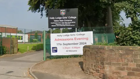 A black sign, behind a sandstone wall, reading 'Holly Lodge Girls' College Visitor Main Entrance'. School building are visible in the background behind a green mesh wire fence. 