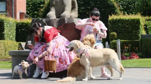 Two women dressed in pink and both with dark hair and sun glasses are with three light-coloured dogs. They are feeding the dogs ice cream