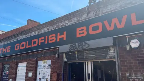 The front facade of a large aquatic store in a city. Above the shops brick wall is a large black sign with orange lettering, reading: "The Goldfish Bowl"