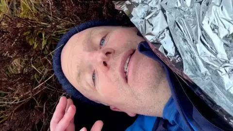 BBC Scotland political editor Glenn Campbell lying on the ground with a blue beanie hat and wrapped in a silver blanket