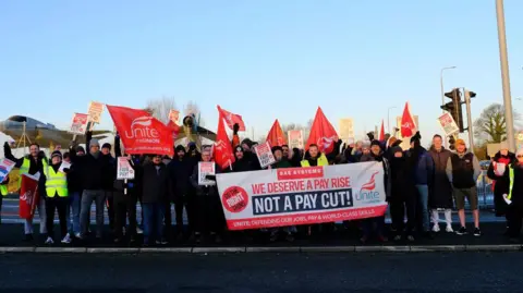 Ross Quinn/Supplied A small group of protesters holding flags and placards pushing for a pay rise. They are stood at the side of a road and dressed warmly in puffer jackets, jeans and woolly hats