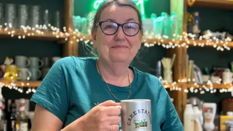 Simon Thake A woman with black rimmed glasses and wearing a green t-shirt stands infront of fairy lights and holds a mug