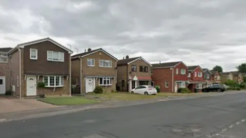 A suburban residential street lined with detached houses on one side under a cloudy, overcast sky. The viewpoint is taken from across a wide road, looking diagonally down the street as it stretches into the distance on the right-hand side.