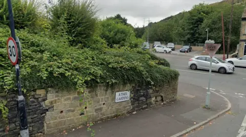 A brick wall in a state of disrepair. A street sign reads Athol Gardens. Beyond is a busy main road