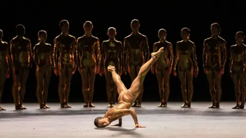 Edinburgh International Festival/Reneff-Olson Productions A dancer is balancing on both arms with their head to the floor and legs extended upwards in a diagonal position, showing their strength and flexibility. They are dressed in a shimmering gold costume that reflects the stage lights.
In the background, a line of performers stands evenly spaced, all wearing matching metallic gold bodysuits. 