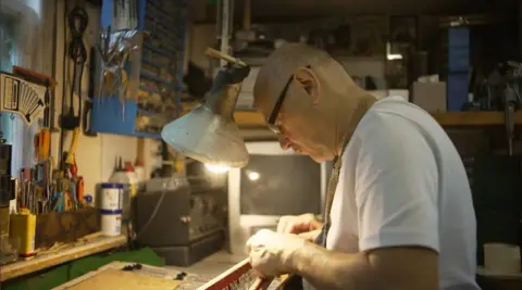 A man, surrounded by tools, sits at a work bench with the inner mechanisms of an accordion in front of him